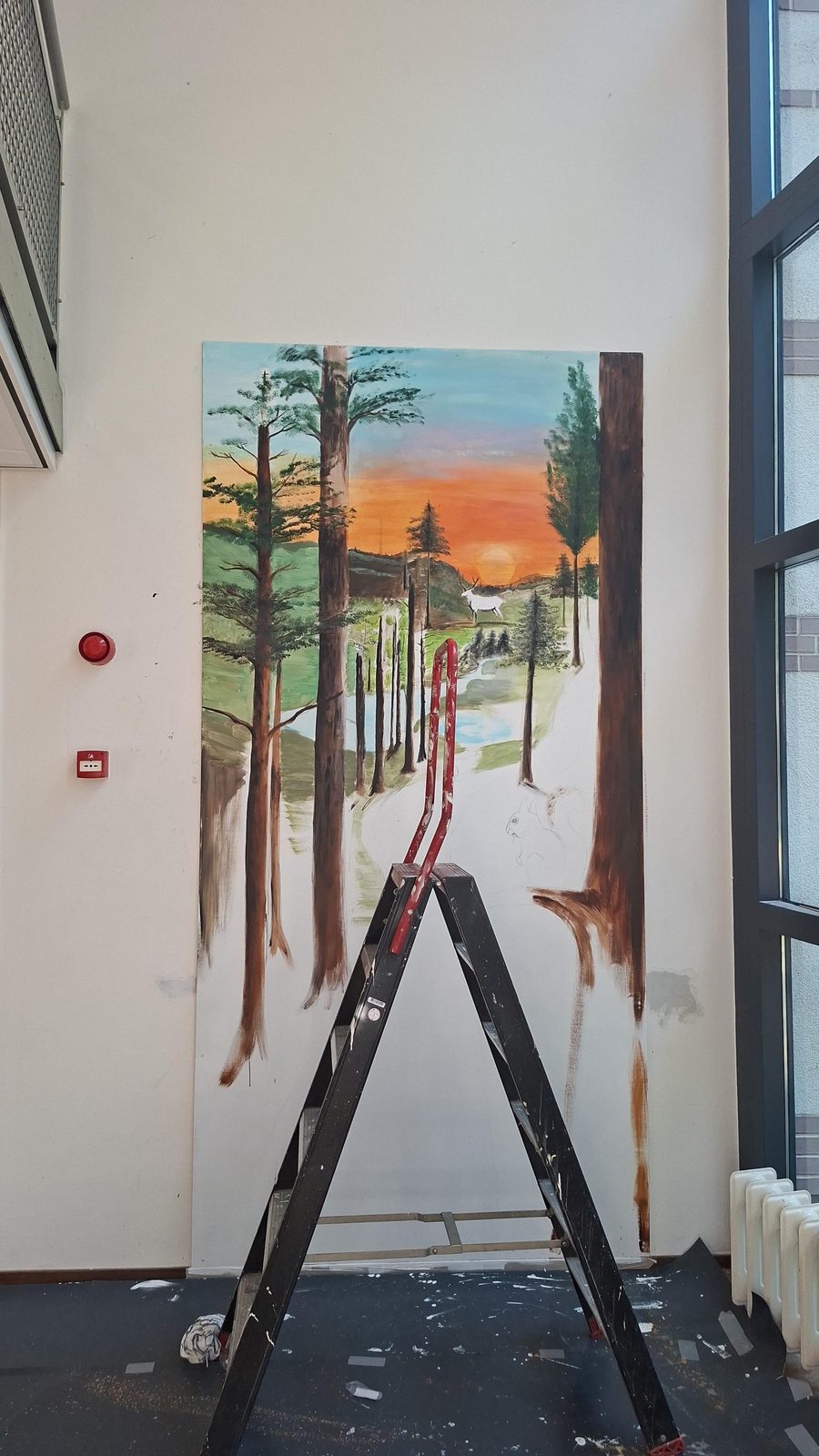 Muurschildering van een dennenbos bij zonsondergang met een hert, ruim drie meter hoog op een wand in een schoolgebouw
